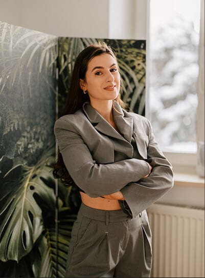 Maria Dziurzyńska stoi z założonymi rękami w stroju służbowym w swoim gabinecie jako dietetyk kliniczny. W tle parawan z roślinami.