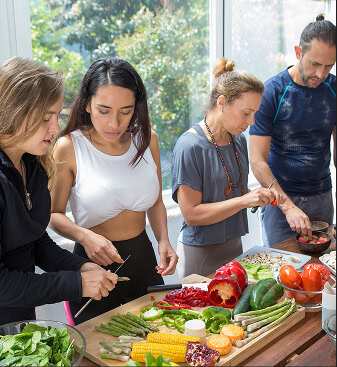 Zdjęcie z warsztatów żywieniowo treningowych, gdzie jest pełno owoców i warzyw na stole a osoby przygotowują sobie jedzenie.